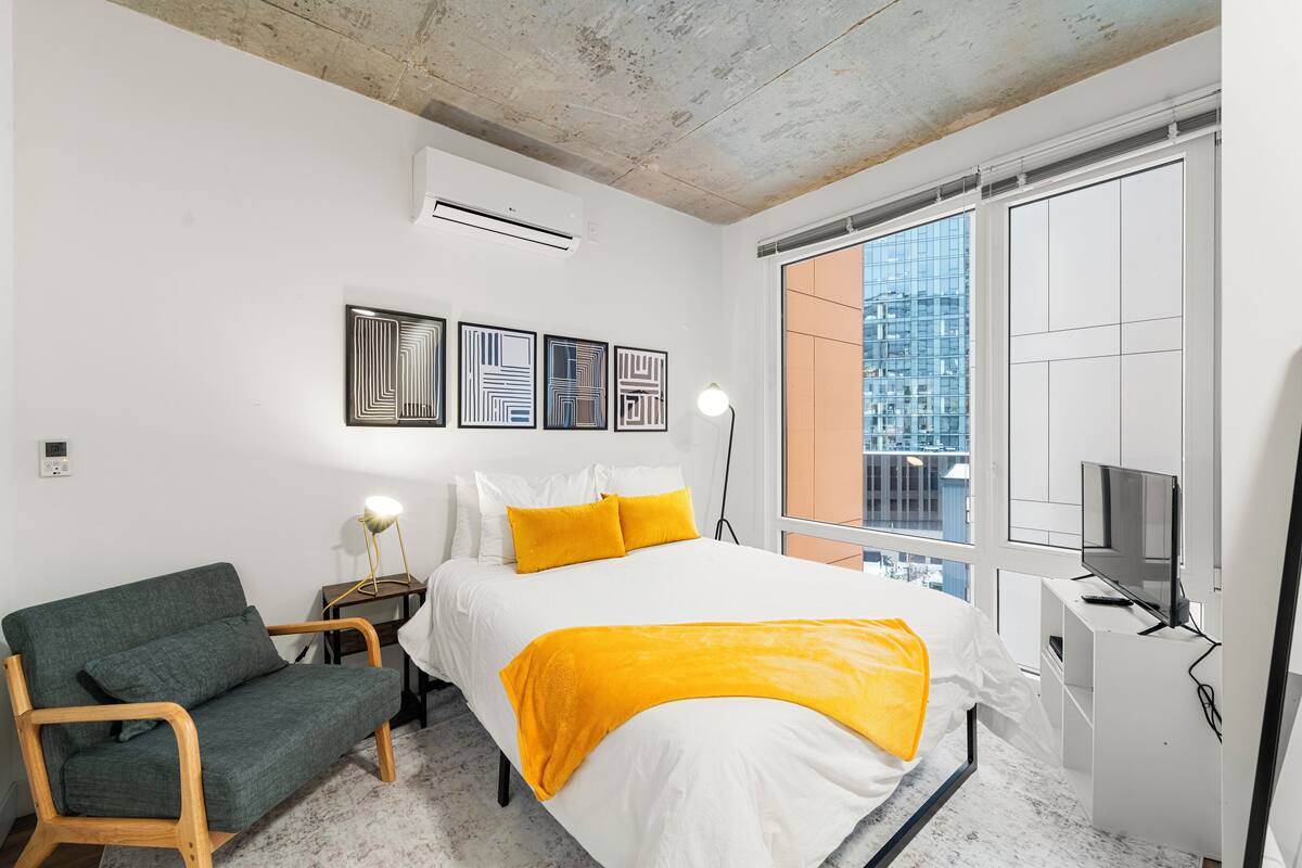 A modern bedroom with a bed, a sofa, a TV, and a window with a city view.