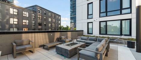 An outdoor terrace with modern furniture, including sofas and a fire pit, surrounded by buildings with large windows and a clear sky.