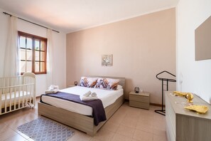 A bedroom with a bed, a crib, a bedside table, a dresser, and a window with curtains. - Holiday Home 'Blue Infinity' with Sea View, Shared Terrace and Air Conditioning (Sant'Isidoro)