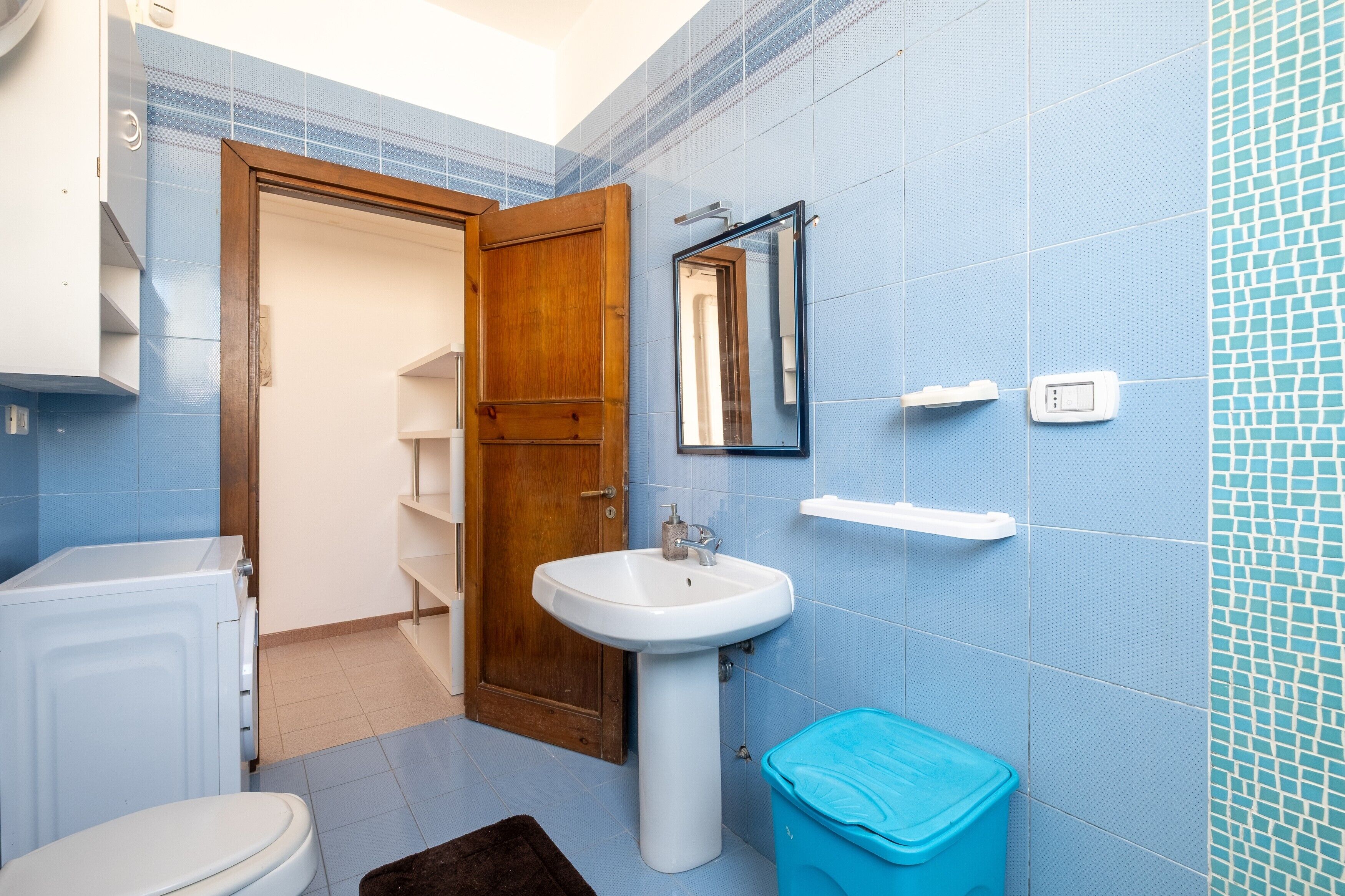 Un bagno con un lavandino bianco, una porta in legno, un cestino della spazzatura blu e un water bianco.
