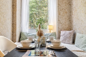 Une table à manger dressée avec des bols, des verres et des couverts, un magazine, et une fenêtre donnant sur un paysage verdoyant.