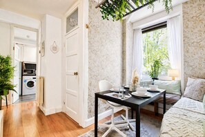 Un espace de vie compact comprenant un coin repas, un canapé, un lave-linge et une vue sur la verdure par une fenêtre.