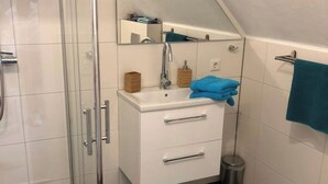 Un baño moderno con cabina de ducha de vidrio, un tocador blanco con dos cajones, un espejo sobre el lavabo y un cubo de basura pequeño.