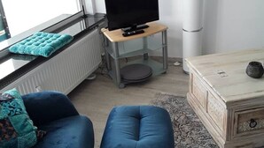 En stue med et fjernsyn på et stativ, en blå sofa, et træskab og en radiator.