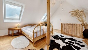 Ein Schlafzimmer mit einem Holzbett, einem runden Teppich, einem Kuhfellkissen, einem kleinen Tisch mit Lampe und einem Fenster mit Blick auf den Himmel.