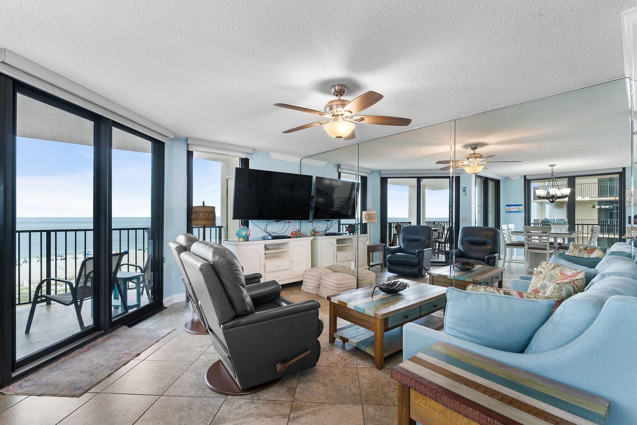 A spacious living room with a large sofa, a recliner, and a coffee table. There is a flat-screen TV mounted on the wall, and a ceiling fan above. The room has large windows offering a view of the beach.