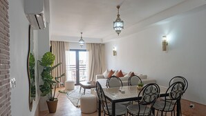 Een moderne woonkamer met een eethoek, met een zithoek, een salontafel en een eettafel met stoelen. Er staat een grote potplant en hangt een lamp.