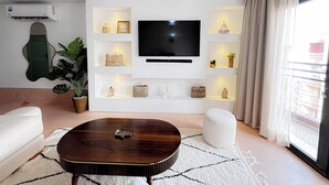 Een moderne woonkamer met een flatscreen televisie, witte planken met decoratieve objecten, een houten salontafel en een potplant.