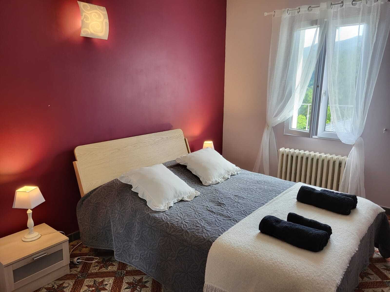 Une chambre à coucher avec un lit, une table de chevet, une fenêtre avec des rideaux légers et un radiateur.