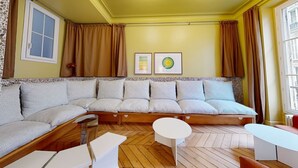 A room with a large L-shaped sofa, a small round table, and a window with curtains. - JOIVY Pleasant 1-bed flat w/ shared courtyard (Paris)