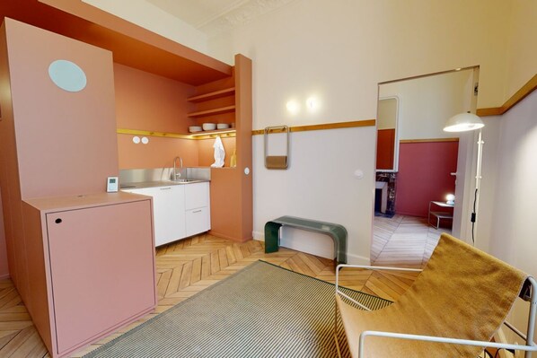 A compact kitchen with a refrigerator, sink, and countertop, a bench, and a mirror reflecting a dining area.