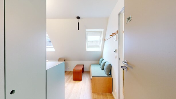 A compact room with a sofa, a small table, a radiator, a window, and a door with a number. - JOIVY 10 Douai 1BR Velux (Paris)