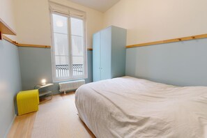 A bedroom with a bed, a radiator, a window, a wardrobe, and a small table with a lamp.