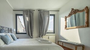 Habitación de hotel con una cama, dos ventanas con cortinas, un banco, un espejo y un radiador.
