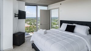 A modern bedroom with a large bed, a TV mounted on the wall, and a city view through the window.