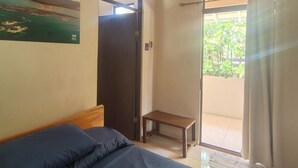 A bedroom with a bed, a small table, a door, and a view of greenery outside.