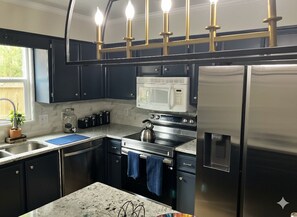 A modern kitchen with dark cabinets, stainless steel appliances, a microwave, oven, and a refrigerator. There are pendant lights above the counter and a window providing natural light. - Golden Isles Retreat near the beach (Brunswick)