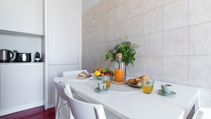 Une cuisine moderne avec des meubles blancs, une table à manger avec des chaises, un panier de fruits, une bouteille de jus d’orange et un vase rempli de fleurs.