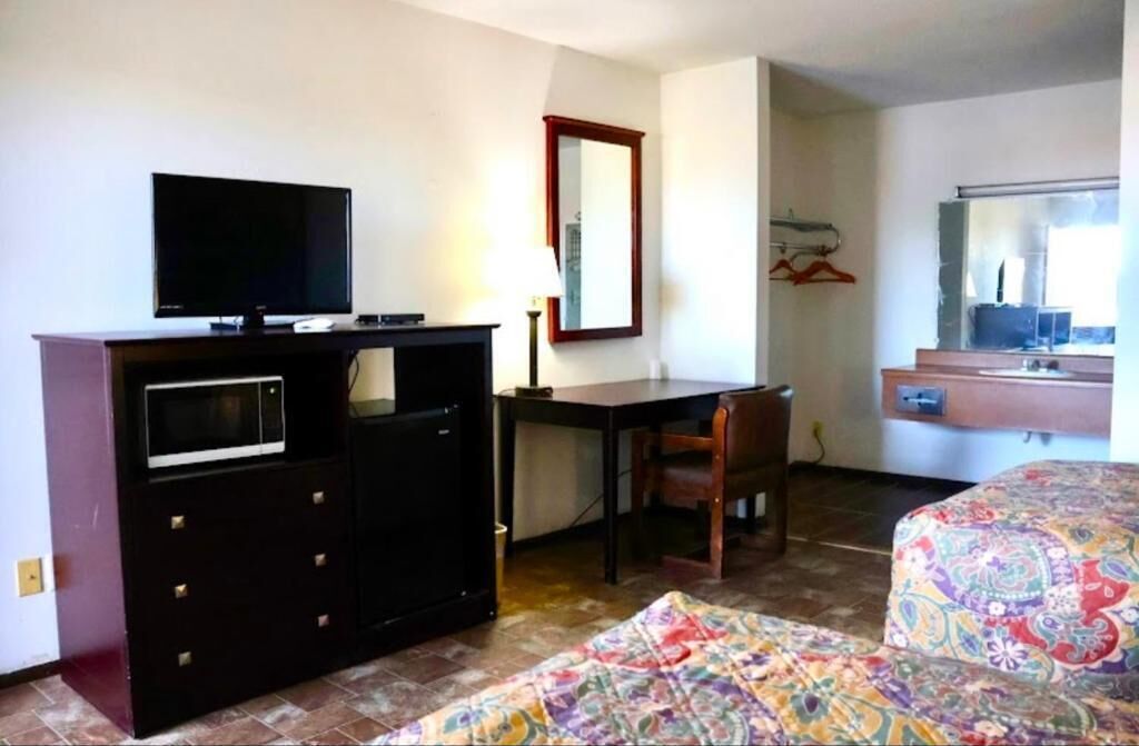 Una habitación de hotel con una cama, un escritorio con televisor, un aparador, una silla y un espejo.