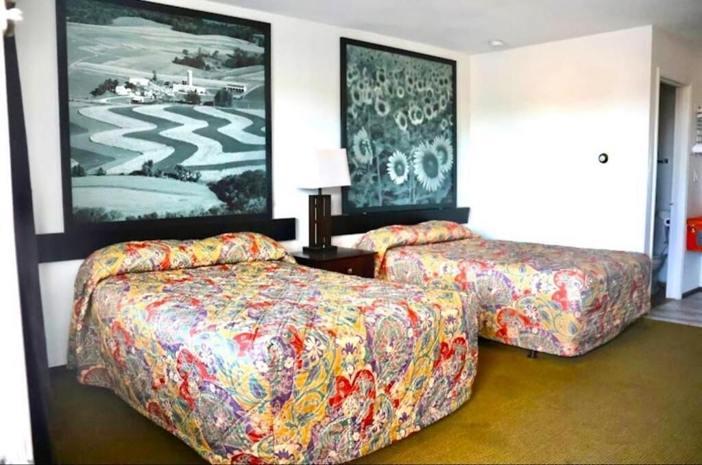 Una habitación de hotel con dos camas, una mesita de noche y dos grandes cuadros con paisajes en las paredes.