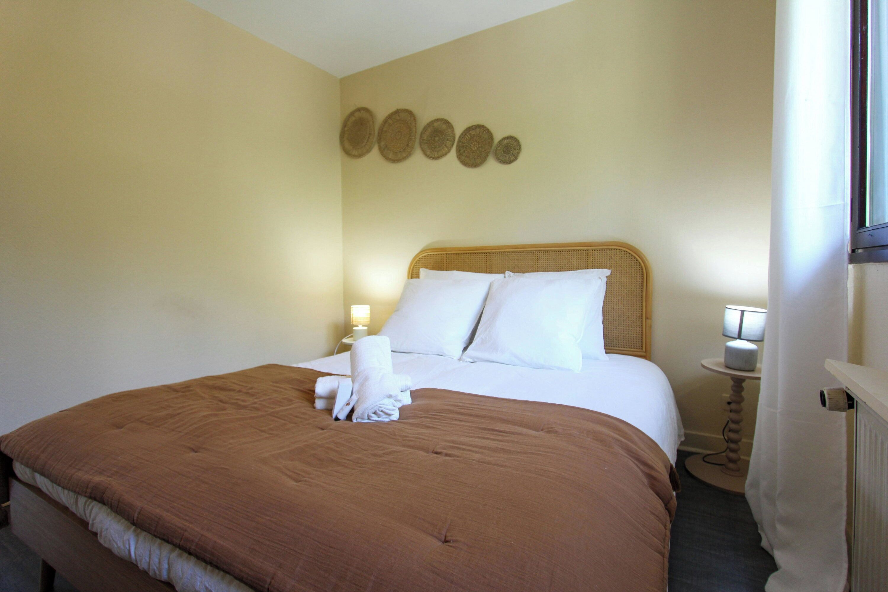 A hotel room with a bed, white linens, a brown bedspread, a wall-mounted lamp, and a small table with a lamp.