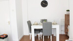 Un comedor con una mesa blanca, dos sillas grises, un reloj negro en la pared y una planta pequeña en la estantería.