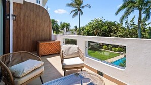 A balcony with wicker chairs, a glass table, and a view of a swimming pool and greenery.