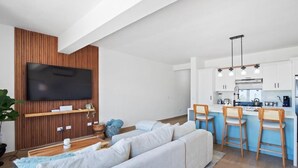 A modern living room with a large flat-screen TV, a sofa, and a kitchen area with bar stools and a blue countertop.