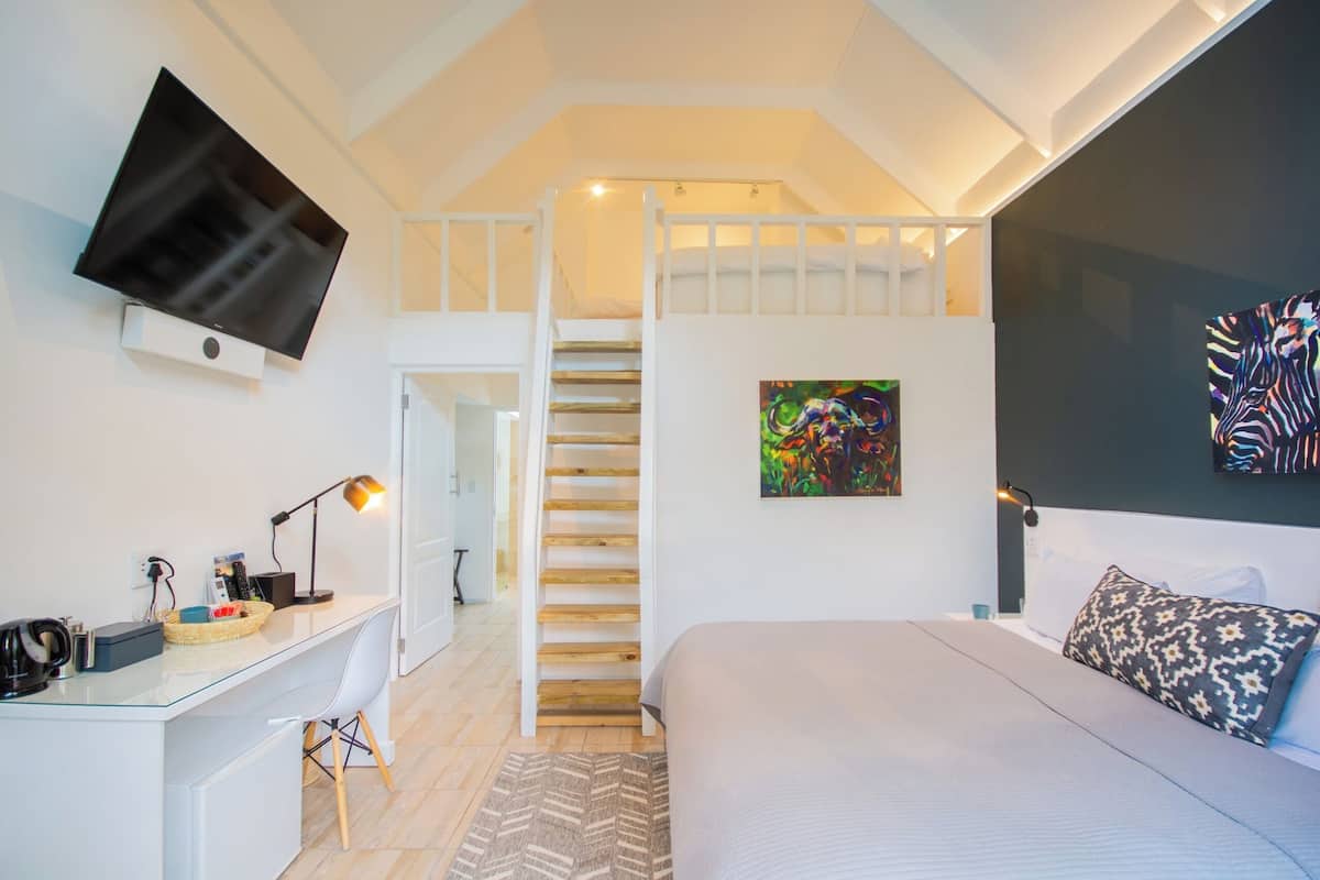 A bedroom with a bed, a desk, a TV mounted on the wall, a staircase, and a painting.