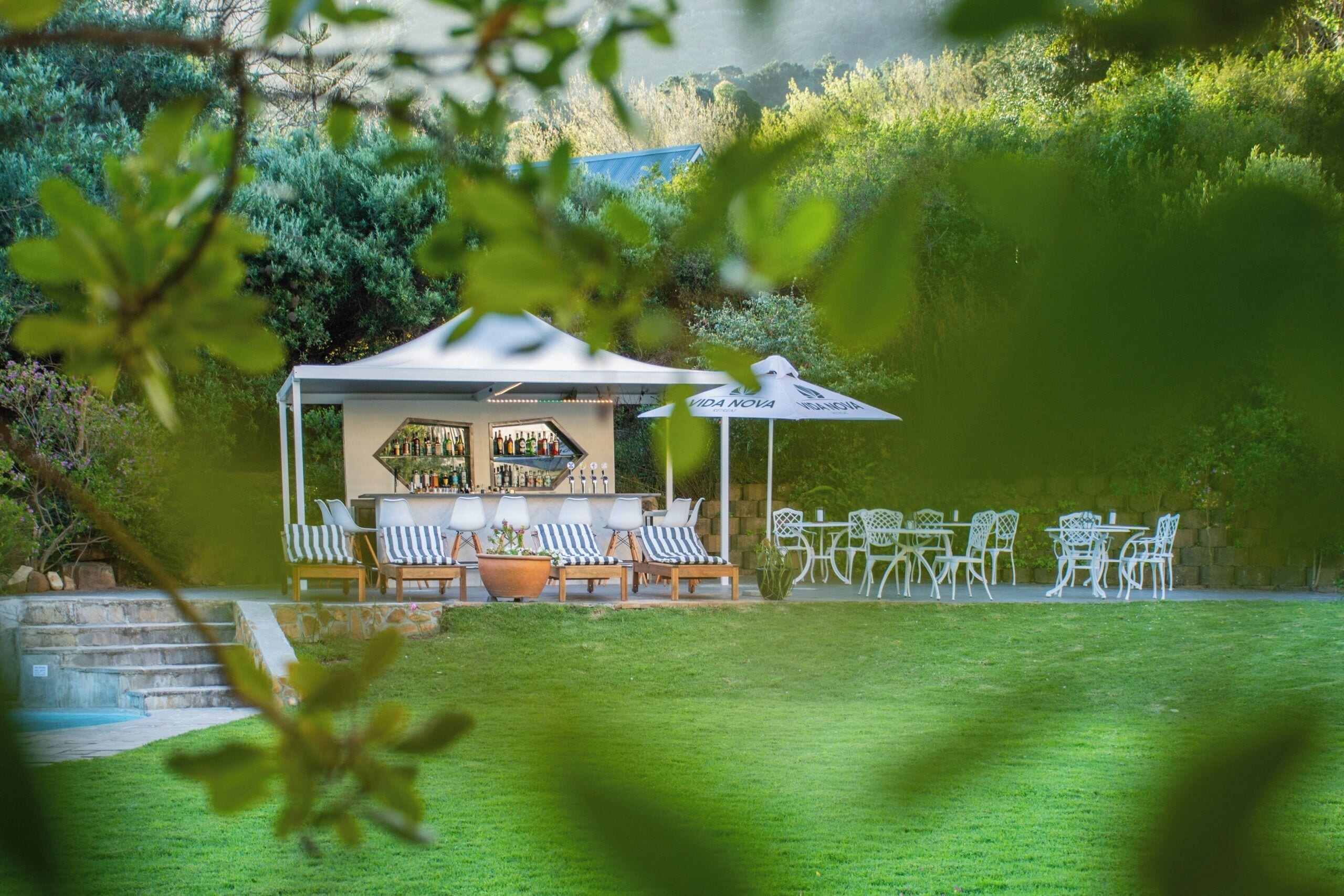 Un espace bar avec des places assises en plein air, un auvent blanc et une vue sur un jardin luxuriant.