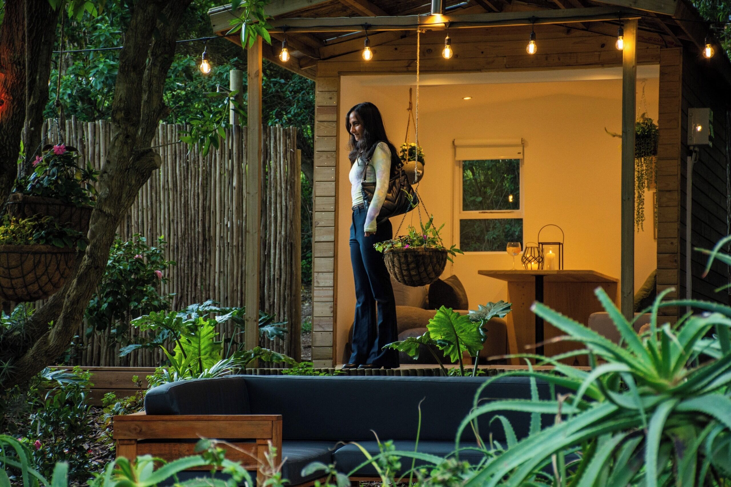 Un espace extérieur accueillant avec un banc en bois, un pot de fleurs suspendu et une table agrémentée d’une bougie.