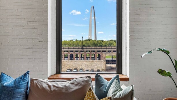 A room with a large window overlooking a bridge and a monument. There are several pillows on a wooden bench in front of the window. - Amazing Riverfront Views - Steps from Arch (St. Louis)