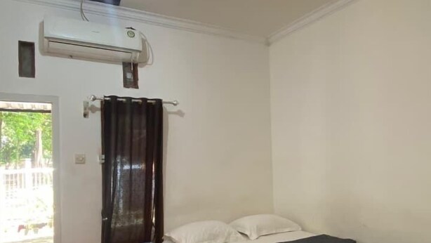 A single bed with a dark mattress and white pillows, a small wooden stool, a wall-mounted air conditioner, and a door leading to an outdoor area. - Backpacker Homestay (Sumberagung)