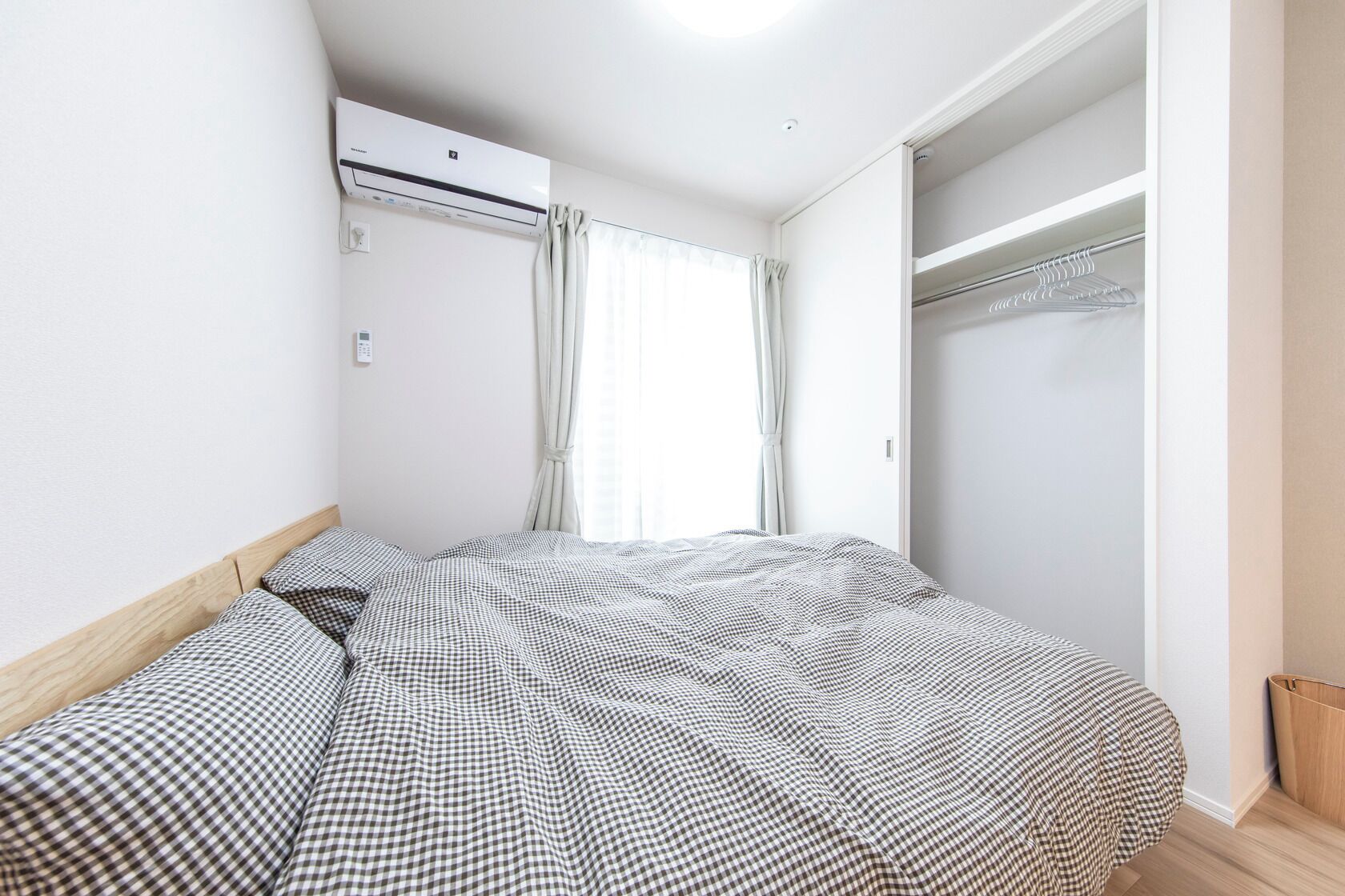 A bedroom with a checkered bedspread, an air conditioning unit, a closet with hanging clothes, and a window with curtains.