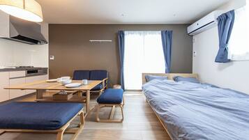 A compact hotel room with a kitchenette, a dining table, two single beds, and a large window with curtains.