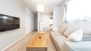 A modern living room with a flat-screen TV, a wooden coffee table, a beige sofa, and a kitchen area in the background. - Logement Aile (Takasaki)