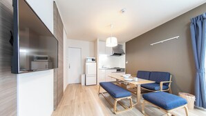 A modern, compact living space with a kitchenette, a dining area with blue upholstered chairs, and a flat-screen TV mounted on the wall. - Logement Aile (Takasaki)
