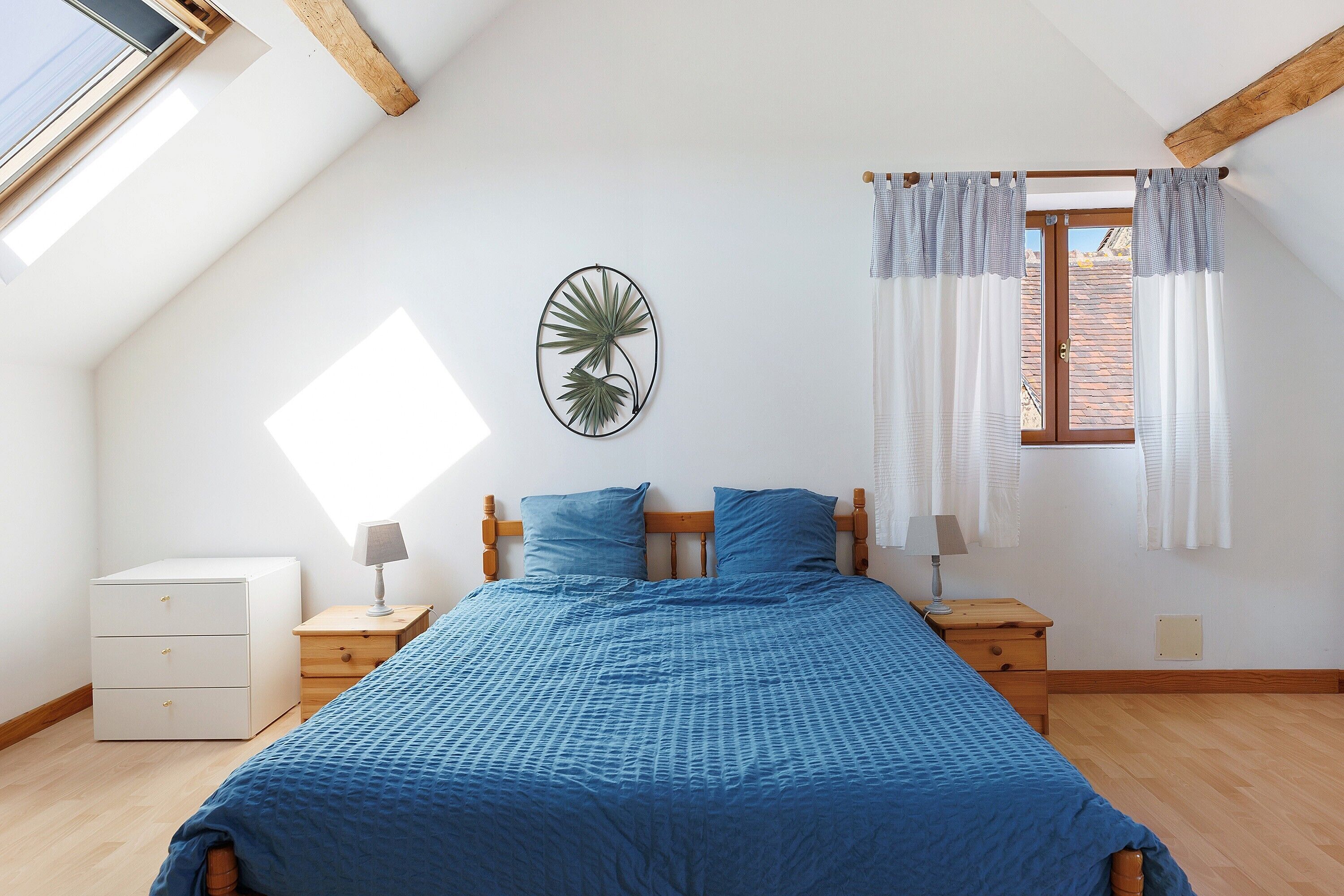 Un dormitorio con techo inclinado, una cama con ropa de cama azul, dos mesitas de noche con lámparas, una planta colgada en la pared y una ventana con cortinas.