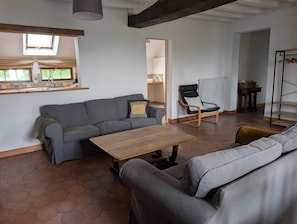 Una sala de estar con un sofá, una mesa de centro de madera, un sillón mecedora y una zona de cocina con ventana y fregadero.