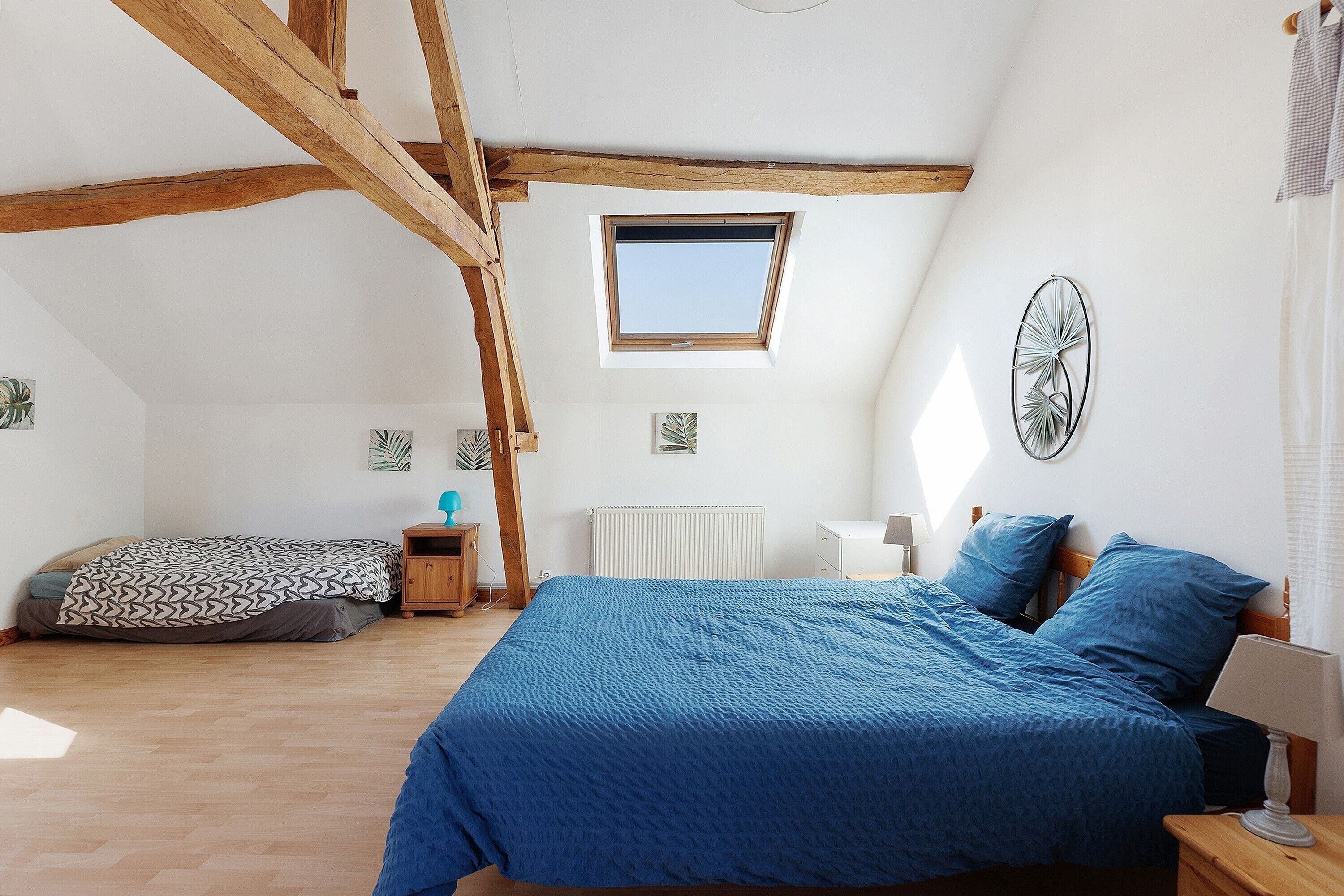 Een slaapkamer met een schuine kap, dakraam, bed met blauw beddengoed, nachtkastje en een ventilator bevestigd aan de muur.