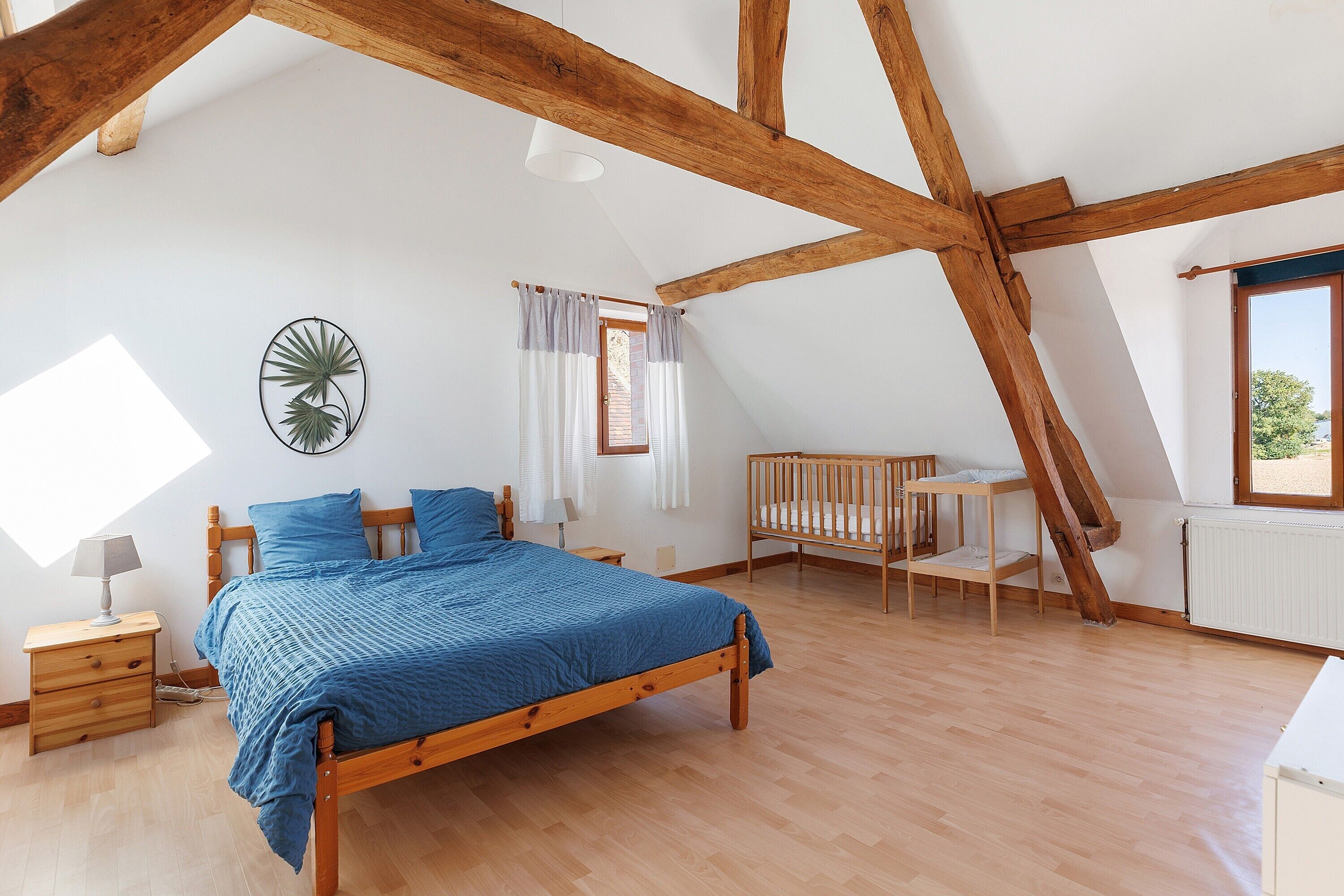 A bedroom with a wooden bed, a crib, a nightstand, a lamp, a wall clock, and a window.