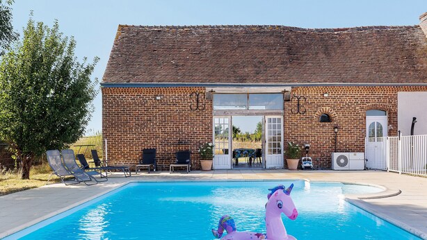 Ein Schwimmbecken mit einem rosa aufblasbaren Einhorn-Schwimmtier, ein Backsteinhaus mit einem Ziegeldach und Sitzgelegenheiten im Freien.