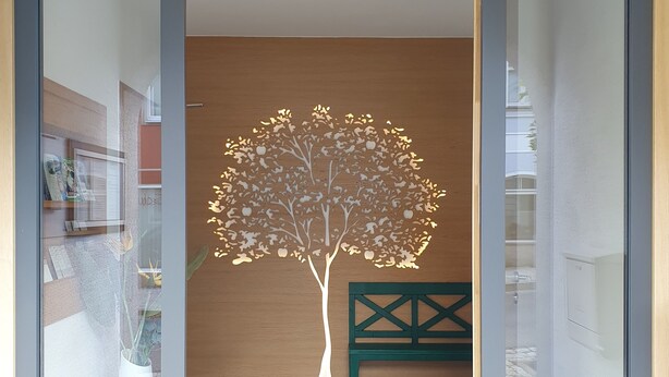 Un intérieur moderne avec une entrée dotée d’une porte en verre, une sculpture d’arbre fixée au mur et un banc vert.