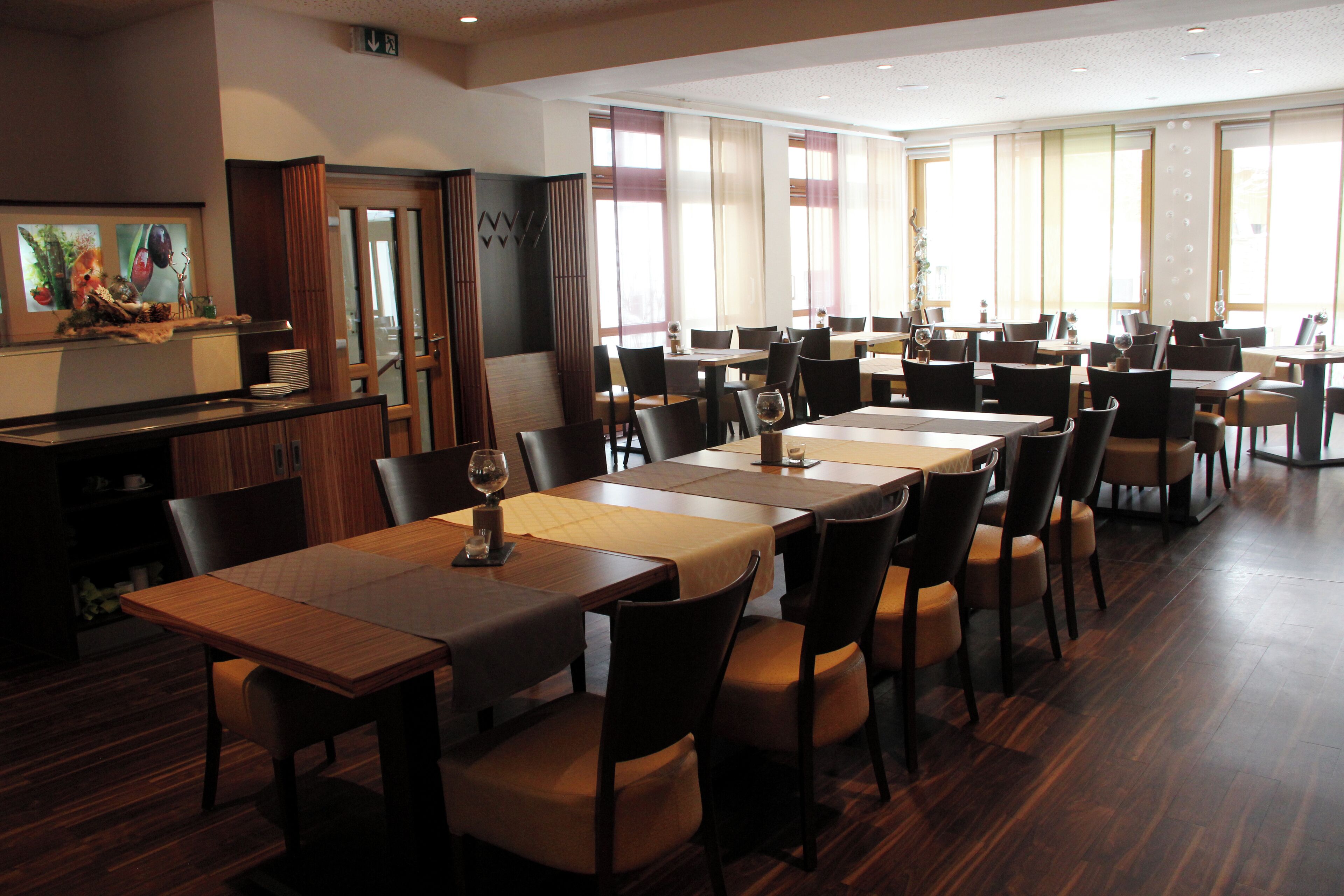 A spacious dining area with multiple tables and chairs, a wooden floor, and large windows allowing natural light.