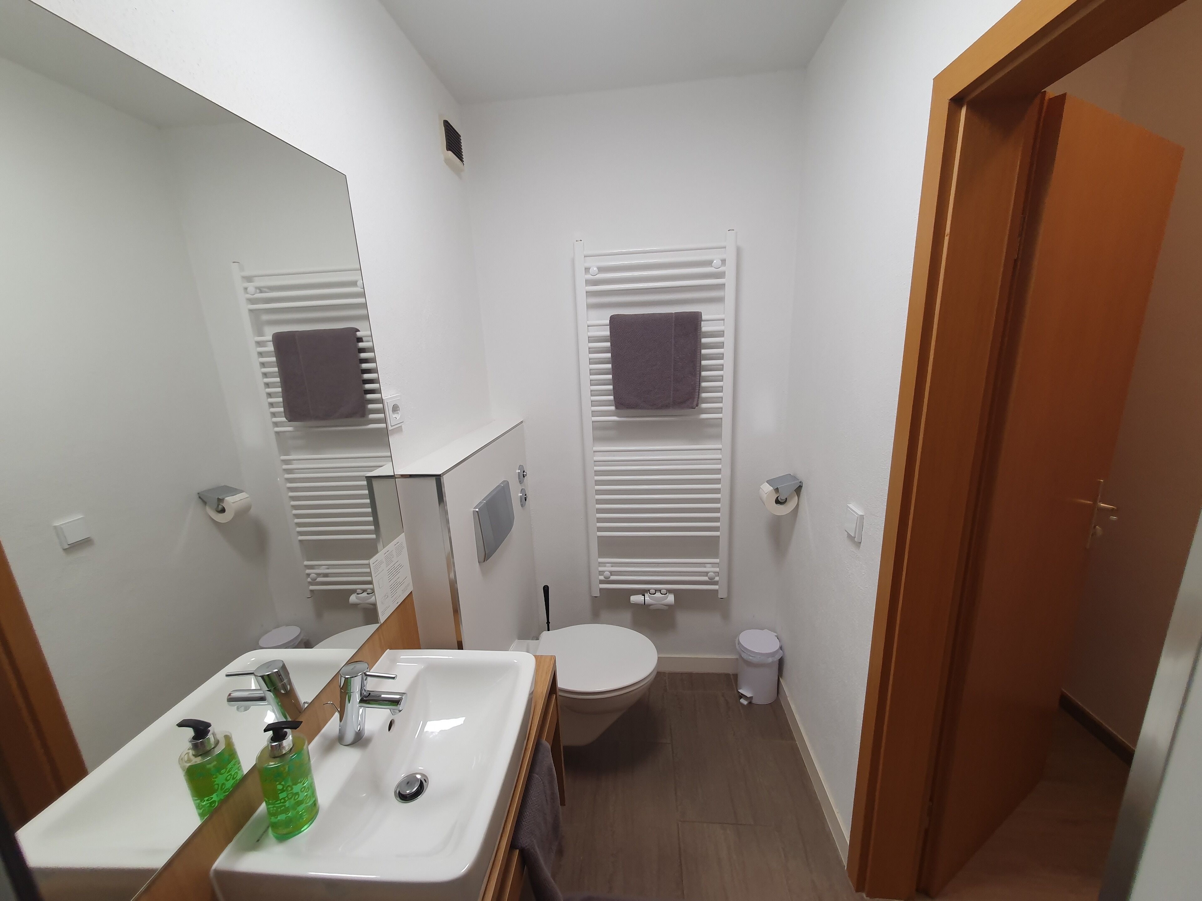 Un baño pequeño con un lavamanos blanco, espejo y inodoro. Hay dos toallas colgadas en toalleros, y se observa un cubo de basura.