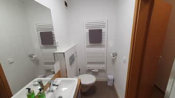 Un baño pequeño con un lavamanos blanco, espejo y inodoro. Hay dos toallas colgadas en toalleros, y se observa un cubo de basura.