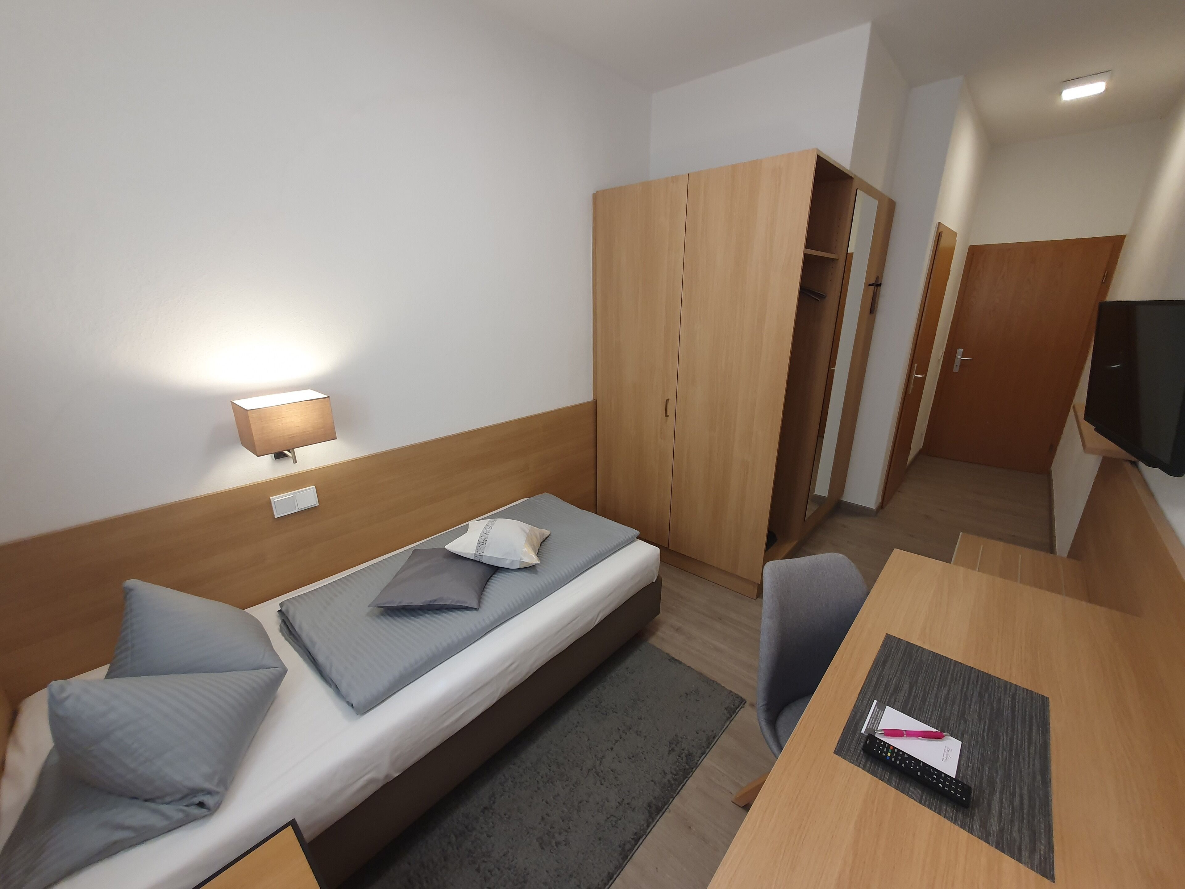 Habitación de hotel pequeña con una cama individual, un escritorio, una silla, un armario y un televisor.