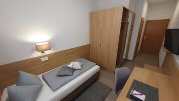 Habitación de hotel pequeña con una cama individual, un escritorio, una silla, un armario y un televisor.