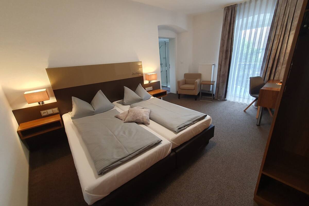 Een hotelkamer met een groot bed, twee nachtkastjes met lampen, een stoel, een bureau en uitzicht op het buitengebied door het raam.