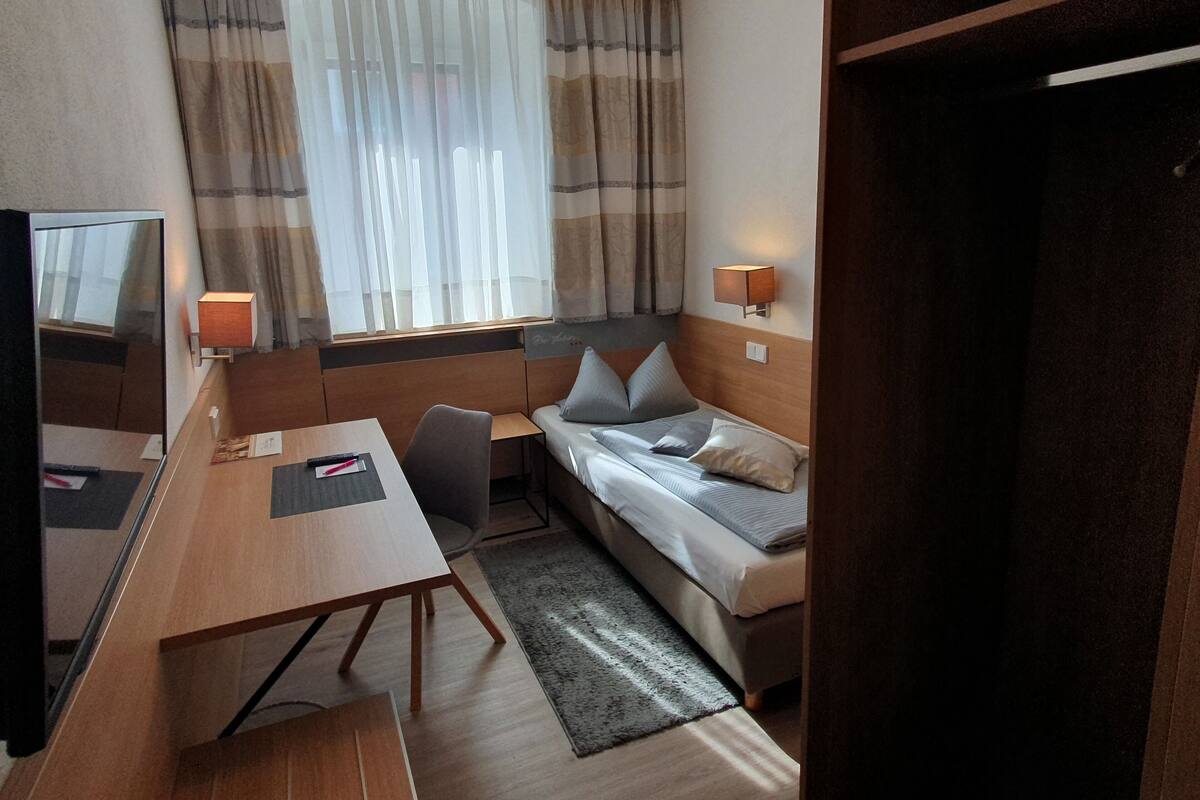 Een kleine hotelkamer met een enkel bed, een bureau, een stoel, een televisie en twee wandlampen.